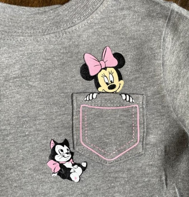 Puede incluir: Una camiseta gris con un bolsillo que presenta a una Minnie Mouse de dibujos animados asom&aacute;ndose por la parte superior y un gato de dibujos animados con un lazo rosa en la parte inferior.
