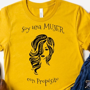 以下が含まれることがあります： マスタードイエローのTシャツに、スペイン語で書かれた言葉に囲まれた女性の顔の黒いグラフィックが描かれています。テキストは「Soy una MUJER con Prop&oacute;sito」です。