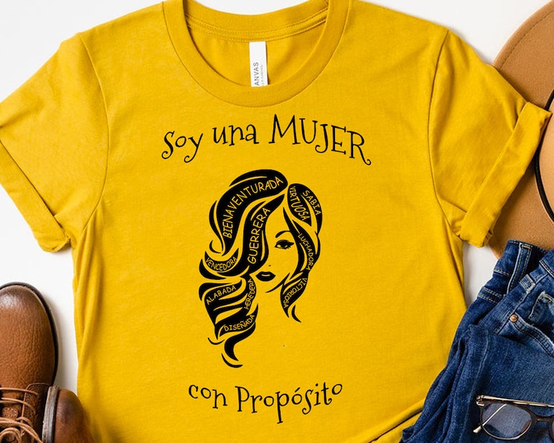 以下が含まれることがあります： マスタードイエローのTシャツに、スペイン語で書かれた言葉に囲まれた女性の顔の黒いグラフィックが描かれています。テキストは「Soy una MUJER con Prop&oacute;sito」です。