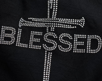 Puede incluir: Una prenda de tela negra con la palabra BLESSED escrita en pedrería transparente y reflectante. Un diseño de cruz vertical, también construido con pequeños brillantes centelleantes, se sitúa detrás de las letras, cruzando la palabra.