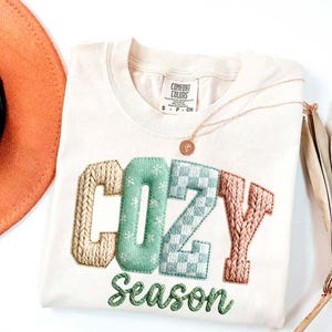 May include: Cream-colored Comfort Colors t-shirt with the words "COZY Season" embroidered in a cozy, textured style. The letters are in beige, mint green, and light blue. A gold necklace is draped across the shirt.