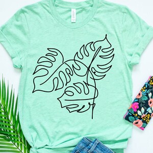 May include: A light green t-shirt with a black line drawing of tropical leaves. The leaves are stylized and resemble monstera plants.