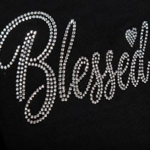 Könnte beinhalten: Eine schwarze Stoffoberfläche mit dem Wort Blessed in einer kursiven Schriftart. Der Schriftzug besteht aus kleinen, klaren, reflektierenden Strasssteinen, die einen funkelnden Effekt auf dem dunklen Hintergrund erzeugen. Ein kleines Herz befindet sich über dem Buchstaben d.