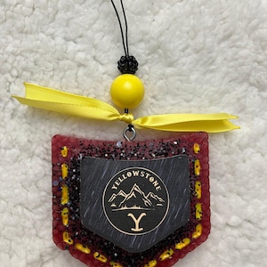 Puede incluir: Un ambientador de coche rojo, amarillo y negro con un logotipo de Yellowstone en blanco y negro. El logotipo presenta una cordillera y la letra Y. El ambientador cuelga de una cinta amarilla.