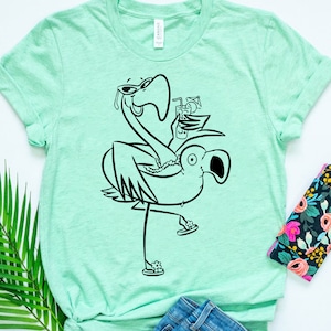 May include: A light green t-shirt with a black and white illustration of a flamingo wearing sunglasses and holding a drink. The flamingo is riding on an inflatable flamingo.