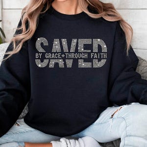 K&ouml;nnte beinhalten: Schwarzes Sweatshirt mit dem Text "SAVED BY GRACE + THROUGH FAITH" in einem Strass-Design. Das Sweatshirt ist ein Rundhals-Stil und wird von einer Person getragen.