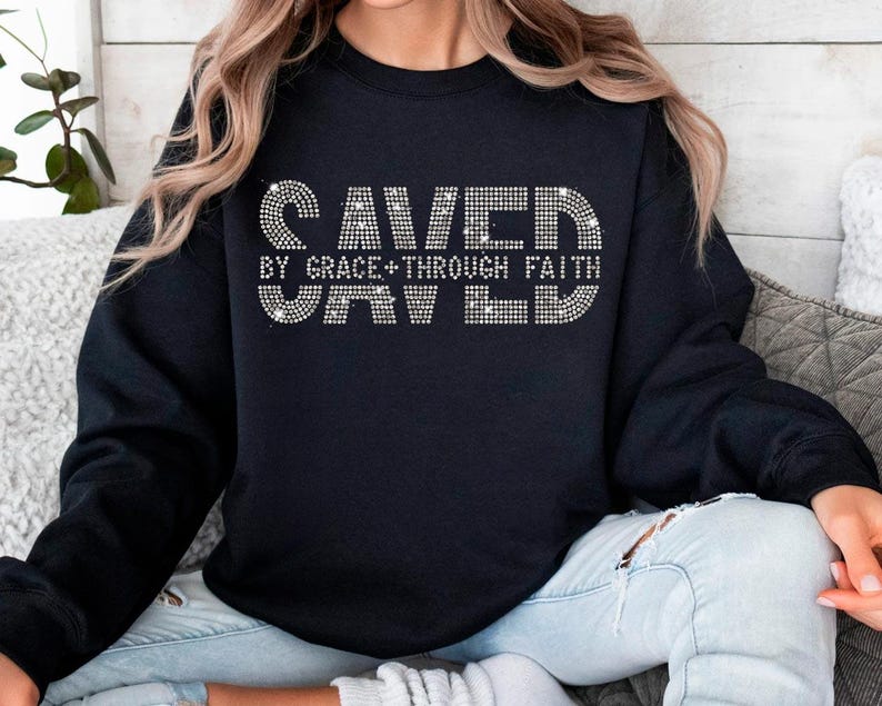 K&ouml;nnte beinhalten: Schwarzes Sweatshirt mit dem Text "SAVED BY GRACE + THROUGH FAITH" in einem Strass-Design. Das Sweatshirt ist ein Rundhals-Stil und wird von einer Person getragen.