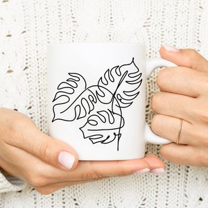 May include: White ceramic mug with a black line drawing of tropical leaves. The mug is being held by a person's hands.