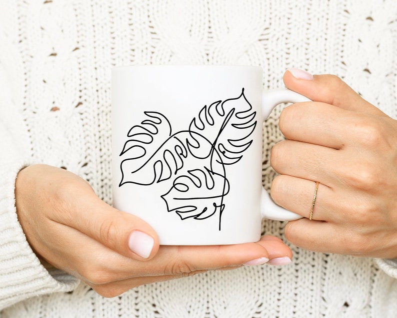 May include: White ceramic mug with a black line drawing of tropical leaves. The mug is being held by a person's hands.