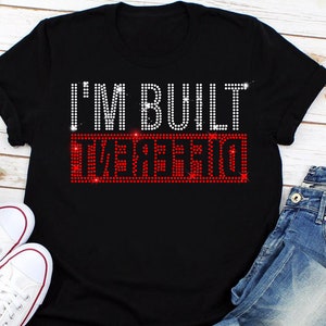 May include: Black t-shirt with the text "I'M BUILT DIFFERENT" in white rhinestones. The word "DIFFERENT" is in red rhinestones.