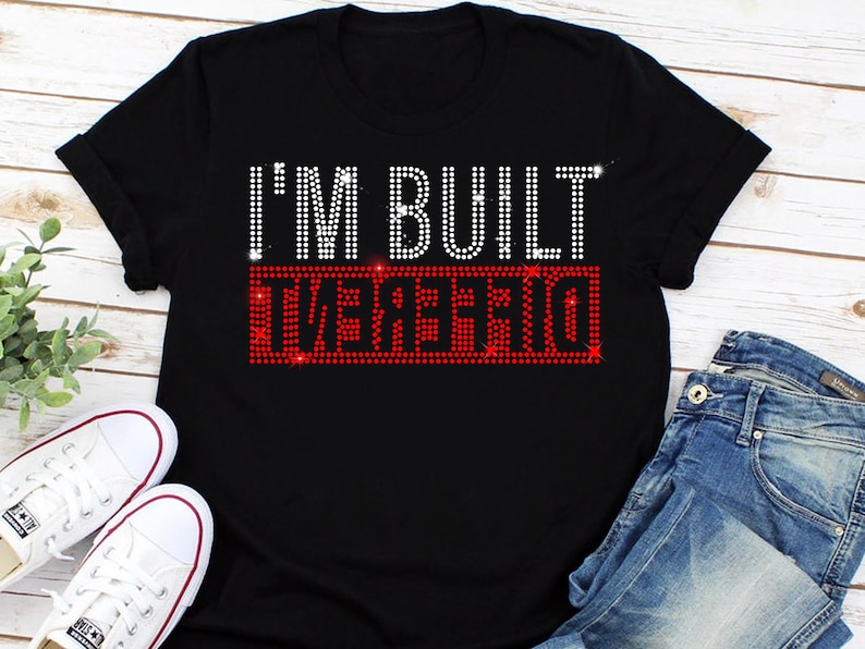 May include: Black t-shirt with the text "I'M BUILT DIFFERENT" in white rhinestones. The word "DIFFERENT" is in red rhinestones.