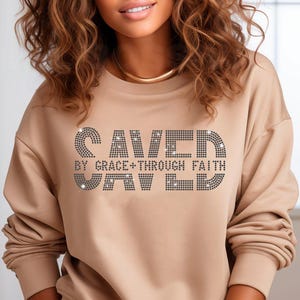 K&ouml;nnte beinhalten: Ein beigefarbenes Sweatshirt mit dem Text "SAVED BY GRACE + THROUGH FAITH" in einem Strass-Design. Das Sweatshirt hat lange &Auml;rmel und einen Rundhalsausschnitt.