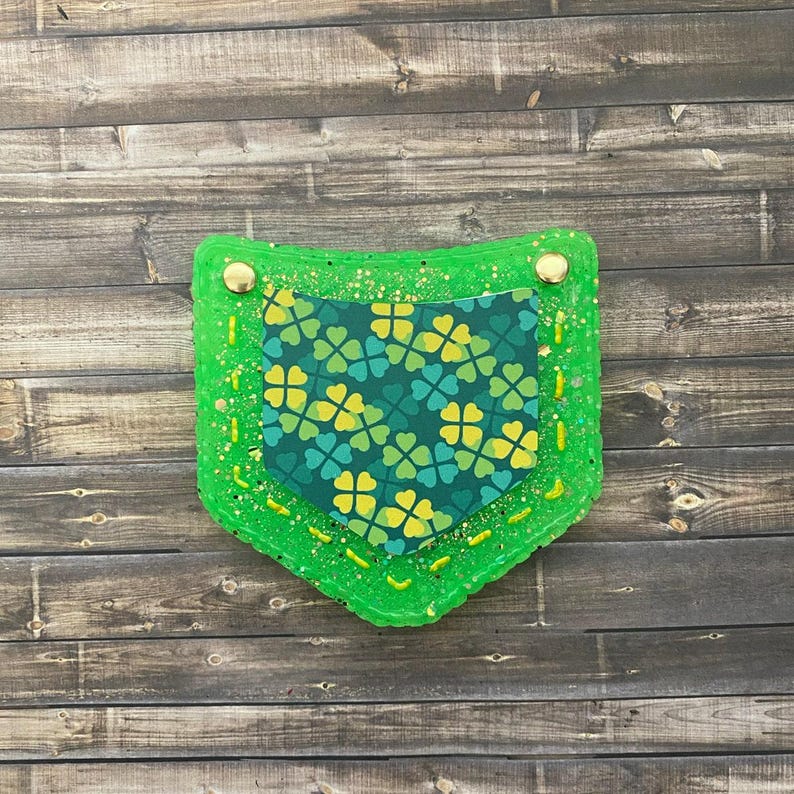 Puede incluir: Un bolsillo de resina verde con purpurina con un patr&oacute;n de tr&eacute;bol en su interior. El bolsillo tiene un bot&oacute;n dorado a cada lado y costuras amarillas alrededor de los bordes.