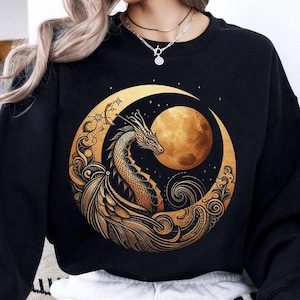 May include: Black sweatshirt with a gold crescent moon design featuring a dragon and a full moon. The design is intricate and detailed, with a swirling pattern around the moon.