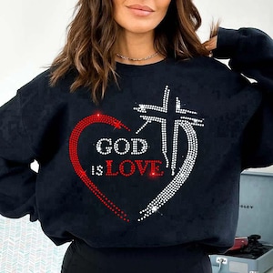 Puede incluir: Sudadera negra con un diseño de pedrería que presenta un corazón rojo y una cruz blanca. Las palabras "GOD is LOVE" están escritas con pedrería dentro del corazón. Una prenda informal de temática religiosa.