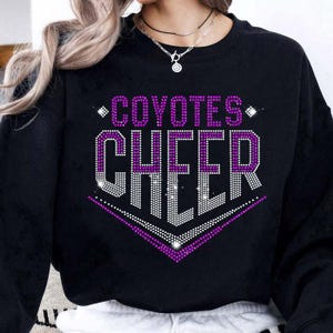 May include: Black sweatshirt with the words "COYOTES CHEER" in a purple and white rhinestone design. The text is arranged in a stacked format, with "COYOTES" above "CHEER". The design includes diamond accents and a chevron shape.