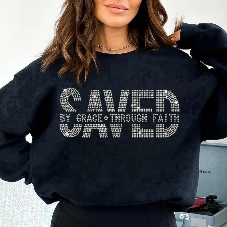 K&ouml;nnte beinhalten: Schwarzes Sweatshirt mit dem Text "SAVED BY GRACE + THROUGH FAITH SAVED" in einem funkelnden, strass&auml;hnlichen Design. Das Sweatshirt ist dunkel und hat einen Rundhalsausschnitt.