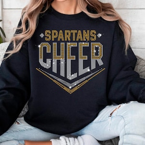 May include: Black sweatshirt with the words "SPARTANS CHEER" in gold and silver rhinestone-like lettering. The design is centered on the chest. The sweatshirt is a crew neck style.