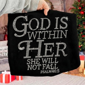 May include: Black tote bag with the text "GOD IS WITHIN HER SHE WILL NOT FALL. PSALM 46:5" in sparkling, rhinestone-like letters. The bag is held up by a person. The background includes a Christmas tree and wrapped gifts.