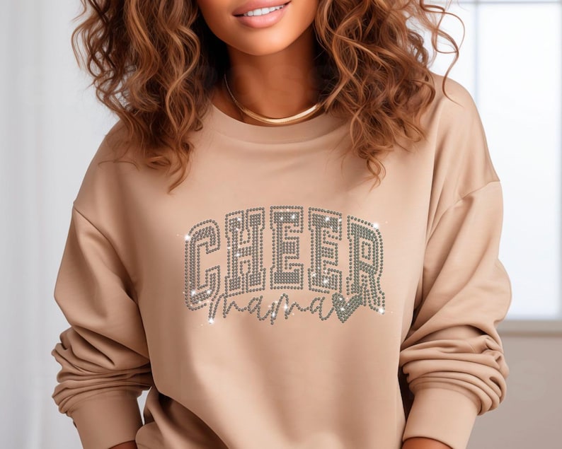 Op de afbeelding: Een beige sweatshirt met de woorden "CHEER mama" in zilveren strass.