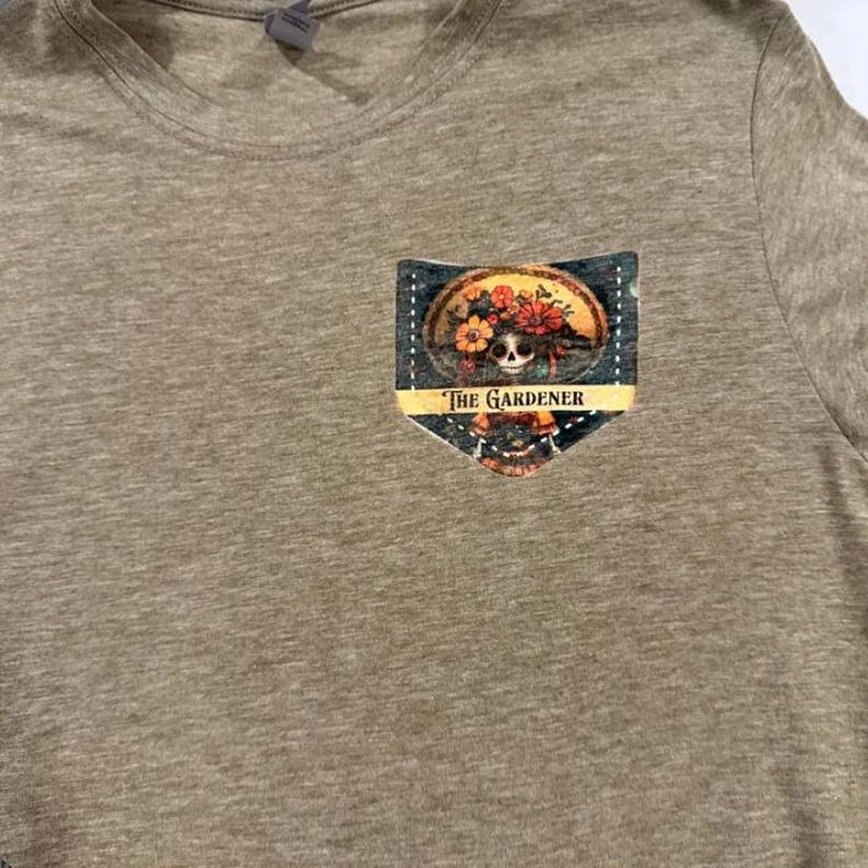 Puede incluir: Una camiseta marr&oacute;n con un bolsillo que presenta un gr&aacute;fico colorido de una calavera con una corona de flores. El texto "The Gardener" est&aacute; impreso debajo de la calavera.
