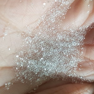 May include: Close-up of a pile of clear, round, glass beads. The beads are small and translucent, and they are scattered on a light-colored surface.