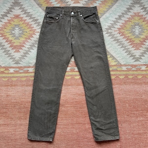 May include: A pair of olive-green denim jeans with a button fly and classic five-pocket design. The jeans are laid flat on a patterned rug. The pants appear to be a straight-leg style.