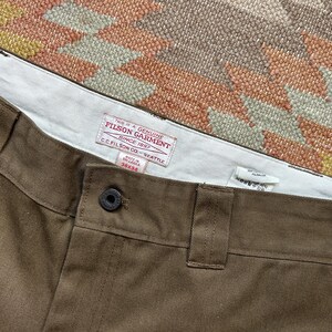 Pode incluir: Close-up de cal&ccedil;as castanhas com fecho de bot&atilde;o e presilhas para cinto. As cal&ccedil;as t&ecirc;m uma cintura interior branca com uma etiqueta vermelha e branca que diz "FILSON GARMENT SINCE 1897". A etiqueta tamb&eacute;m inclui o texto "C.C. FILSON CO. - SEATTLE" e o tamanho "38x34".