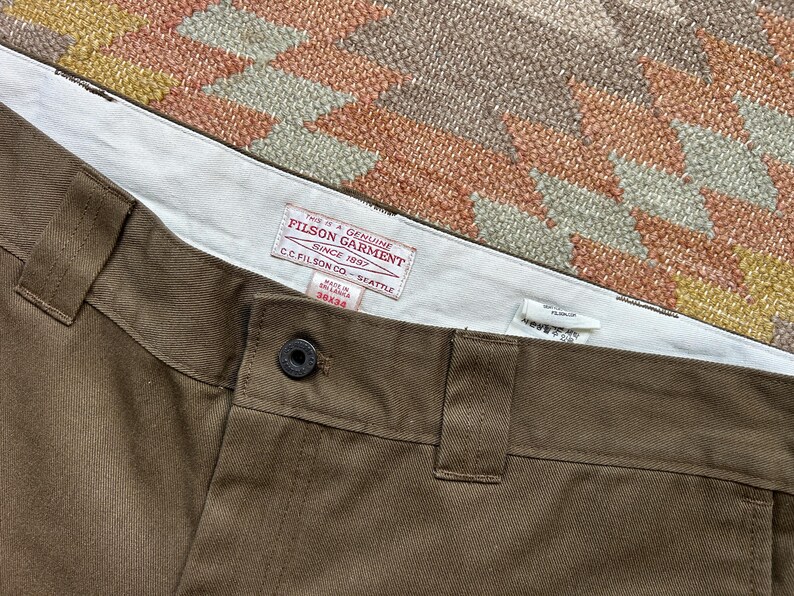 Pode incluir: Close-up de cal&ccedil;as castanhas com fecho de bot&atilde;o e presilhas para cinto. As cal&ccedil;as t&ecirc;m uma cintura interior branca com uma etiqueta vermelha e branca que diz "FILSON GARMENT SINCE 1897". A etiqueta tamb&eacute;m inclui o texto "C.C. FILSON CO. - SEATTLE" e o tamanho "38x34".