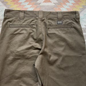 Pode incluir: Close-up da parte de tr&aacute;s de cal&ccedil;as castanhas com presilhas e bolsos. As cal&ccedil;as t&ecirc;m uma pequena etiqueta retangular que diz "FILSON". O tecido tem um ligeiro brilho.
