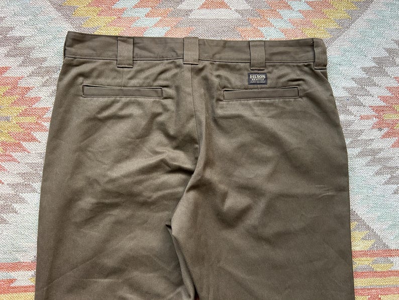 Pode incluir: Close-up da parte de tr&aacute;s de cal&ccedil;as castanhas com presilhas e bolsos. As cal&ccedil;as t&ecirc;m uma pequena etiqueta retangular que diz "FILSON". O tecido tem um ligeiro brilho.