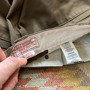 Pode incluir: Close-up de cal&ccedil;as verde-oliva com uma etiqueta vis&iacute;vel. A etiqueta diz "FILSON GARMENT SINCE 1897 SEATTLE" e indica um tamanho de 96.5x86.4 cm. As cal&ccedil;as s&atilde;o feitas no Sri Lanka.