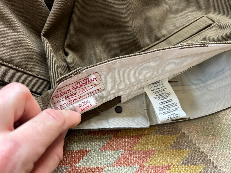 Pode incluir: Close-up de cal&ccedil;as verde-oliva com uma etiqueta vis&iacute;vel. A etiqueta diz "FILSON GARMENT SINCE 1897 SEATTLE" e indica um tamanho de 96.5x86.4 cm. As cal&ccedil;as s&atilde;o feitas no Sri Lanka.