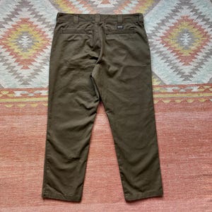 Pode incluir: Cal&ccedil;as verde-oliva com um design cl&aacute;ssico de perna reta. As cal&ccedil;as t&ecirc;m fecho de bot&atilde;o e z&iacute;per, presilhas para cinto e bolsos traseiros. Feitas de tecido dur&aacute;vel, s&atilde;o adequadas para uso di&aacute;rio.