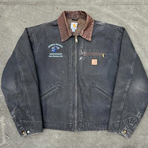 May include: A black Carhartt work jacket with a brown collar and a zipper closure. The jacket features the text "SIERRA PACIFIC INDUSTRIES" in blue and white on the left chest. A small Carhartt logo patch is on the right chest.