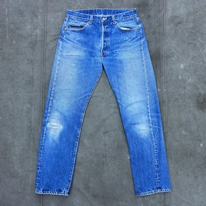 May include: Blue denim jeans with a button fly and classic five-pocket design. The jeans show signs of wear with faded areas and a distressed knee. The jeans are laid flat on a gray surface.