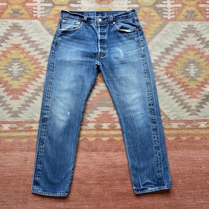 May include: Faded blue denim jeans with a button fly. The jeans show signs of wear and fading, with a classic five-pocket design. The jeans are laid flat, showcasing the details of the denim.