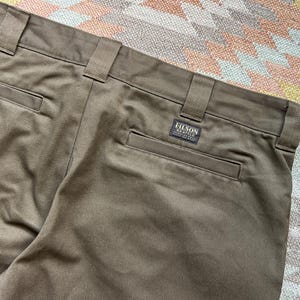 Pode incluir: Close-up de cal&ccedil;as verde-oliva com presilhas e bolsos traseiros. Uma pequena etiqueta retangular com o texto "FILSON SEATTLE" est&aacute; costurada no bolso traseiro. As cal&ccedil;as s&atilde;o feitas de um tecido dur&aacute;vel.