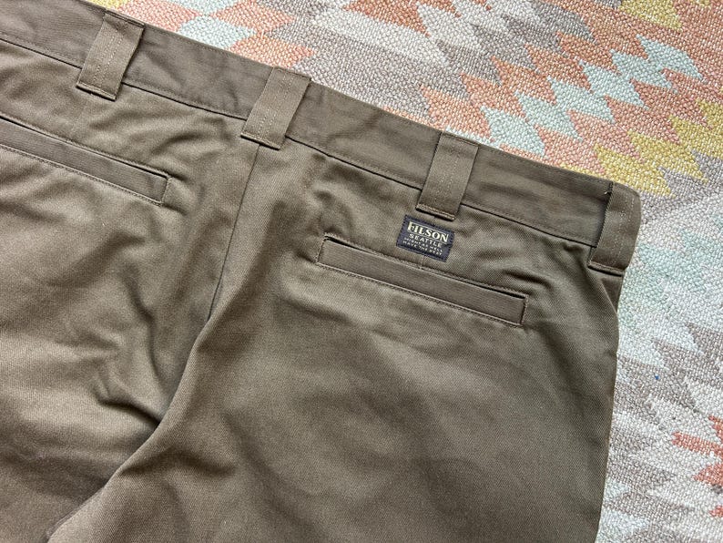 Pode incluir: Close-up de cal&ccedil;as verde-oliva com presilhas e bolsos traseiros. Uma pequena etiqueta retangular com o texto "FILSON SEATTLE" est&aacute; costurada no bolso traseiro. As cal&ccedil;as s&atilde;o feitas de um tecido dur&aacute;vel.
