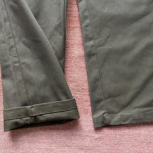 Pode incluir: Close-up de cal&ccedil;as verde-oliva com punhos dobrados. O tecido parece ser um material resistente, e as cal&ccedil;as est&atilde;o bem passadas. O fundo &eacute; uma superf&iacute;cie texturizada, rosada-avermelhada.