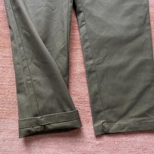 Pode incluir: Close-up de cal&ccedil;as verde-oliva com punhos dobrados. As cal&ccedil;as s&atilde;o feitas de um tecido de cor s&oacute;lida e parecem ser de estilo casual. O fundo &eacute; uma superf&iacute;cie rosa texturizada.