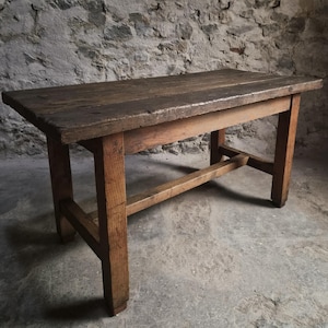 Op de afbeelding: Een rechthoekige houten tafel met een donker, verweerd blad en een lichter bruin onderstel. De tafel heeft vier stevige poten die verbonden zijn door een centrale horizontale steunbalk, met een ruwe textuur en solide bouw.