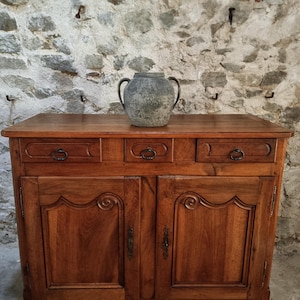 Könnte beinhalten: Ein hölzernes Sideboard mit drei oberen Schubladen mit Metallringgriffen und zwei unteren Schranktüren mit geschnitzten Paneelen und Metallbeschlägen. Ein verwittertes, graues Keramikgefäß mit zwei Henkeln steht mittig auf der Holzoberfläche.