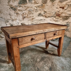 Op de afbeelding: Een rustieke houten tafel met twee lades en een rechthoekig blad. De tafel is gemaakt van lichtbruin hout en heeft een verweerde afwerking. De lades hebben ronde houten knoppen. De tafel wordt ondersteund door vier stevige poten.