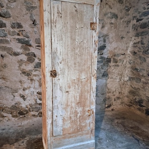 Könnte beinhalten: Ein hoher, schmaler, antiker Holzschrank mit einer einzigen Tür. Der Schrank ist in einem verwitterten Weiß und Hellblau gestrichen, wodurch die Holzmaserung sichtbar wird. Er hat ein schlichtes Design mit einem kleinen Knopf und Scharnieren. Der Schrank steht vor einer Steinmauer.