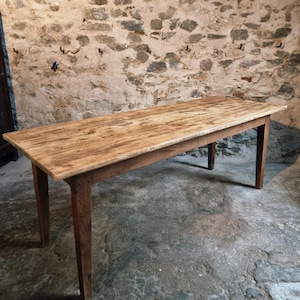Op de afbeelding: Een rechthoekige houten tafel met een lichtbruin tafelblad en vier stevige poten. De tafel heeft een rustieke, verweerde uitstraling, wat suggereert dat het een antiek of vintage stuk is. De tafel is ongeveer 183 cm lang.