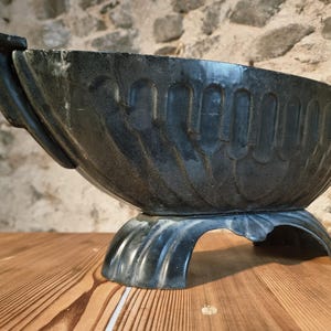 Op de afbeelding: Een donkergrijze, sierlijke plantenbak met een brede, ovale bak en een decoratieve voet. De plantenbak heeft een textuur met verticale groeven en een gebogen, geschulpte voet. De plantenbak is gemaakt van donkergrijs materiaal.