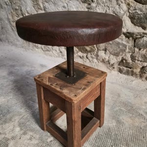 Könnte beinhalten: Ein Hocker mit einer runden, dunkelbraunen Sitzfläche und einem quadratischen Holzgestell. Die Sitzfläche ist über einen zentralen Metallschraubmechanismus mit dem Gestell verbunden, der eine Höhenverstellung ermöglicht. Das Holzgestell hat vier Beine.