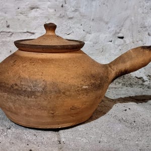 Könnte beinhalten: Eine handgefertigte Teekanne aus Steingut mit Deckel und Ausguss. Die Teekanne hat eine warme, erdige braune Farbe mit dunkleren braunen Akzenten und einer strukturierten Oberfläche. Der Griff ist gebogen und erstreckt sich von der Seite der Kanne.
