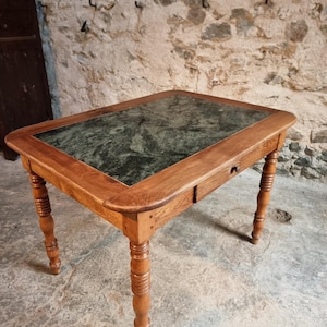 Peut inclure: Une table rectangulaire en bois avec un dessus en marbre vert et un seul tiroir. La table a quatre pieds tournés et est en bois brun clair. La table mesure environ 76 cm de long et 51 cm de large.